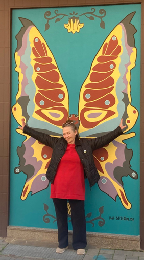 Vor dem farbenfrohen Gemälde eines Schmetterlings, das stilistisch an Art Deco erinnert, steht Isabel Wienold und breitet ihre Arme aus, wie der Schmetterling seine Flügel.