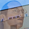 Eine lädierte männliche Schaufensterpuppe mit gesprungenem Brillenglas mischt sich mit der Spiegelung im Fenster des Hauses gegenüber.